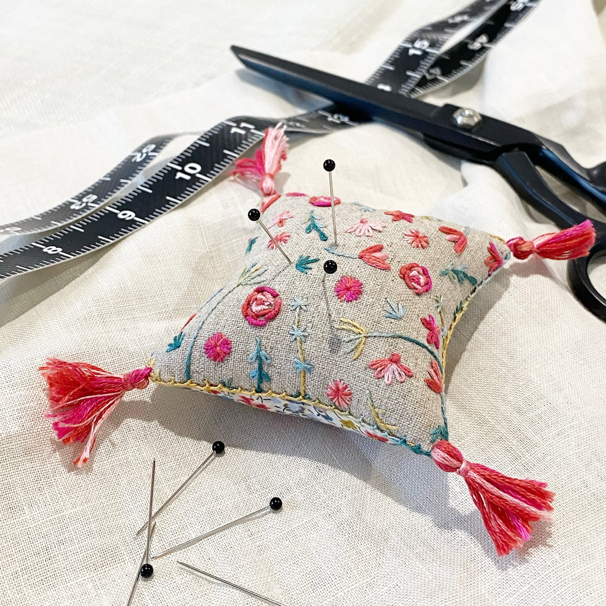 Decorative pincushion with floral embroidery and tassels on a white fabric background with scissors and pins.