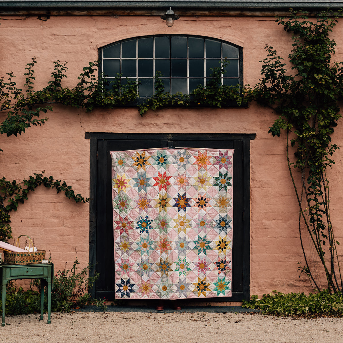 Quilt with floral pattern displayed on a wooden frame against a pink wall with greenery.