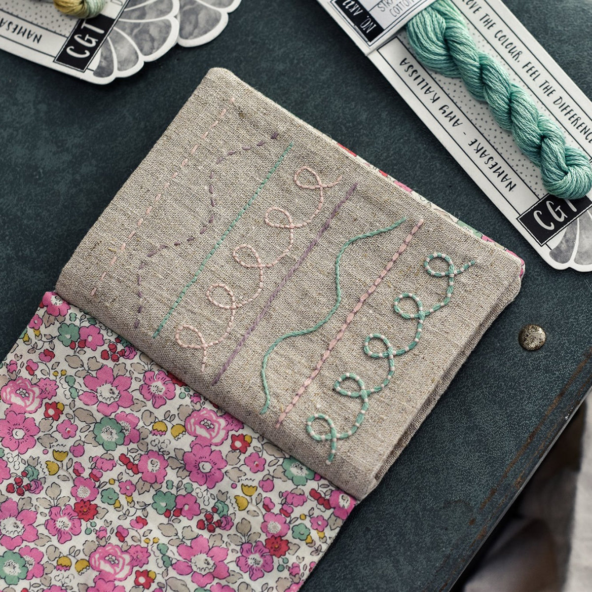 Embroidered fabric with floral pattern and stitch samples, surrounded by embroidery supplies on a dark surface.